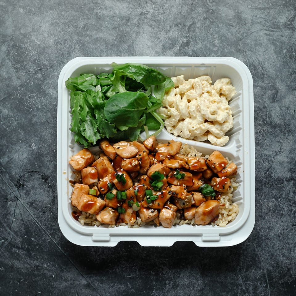 Takeout container with teriyaki chicken, rice, green salad, and macaroni salad on a dark stone surface.