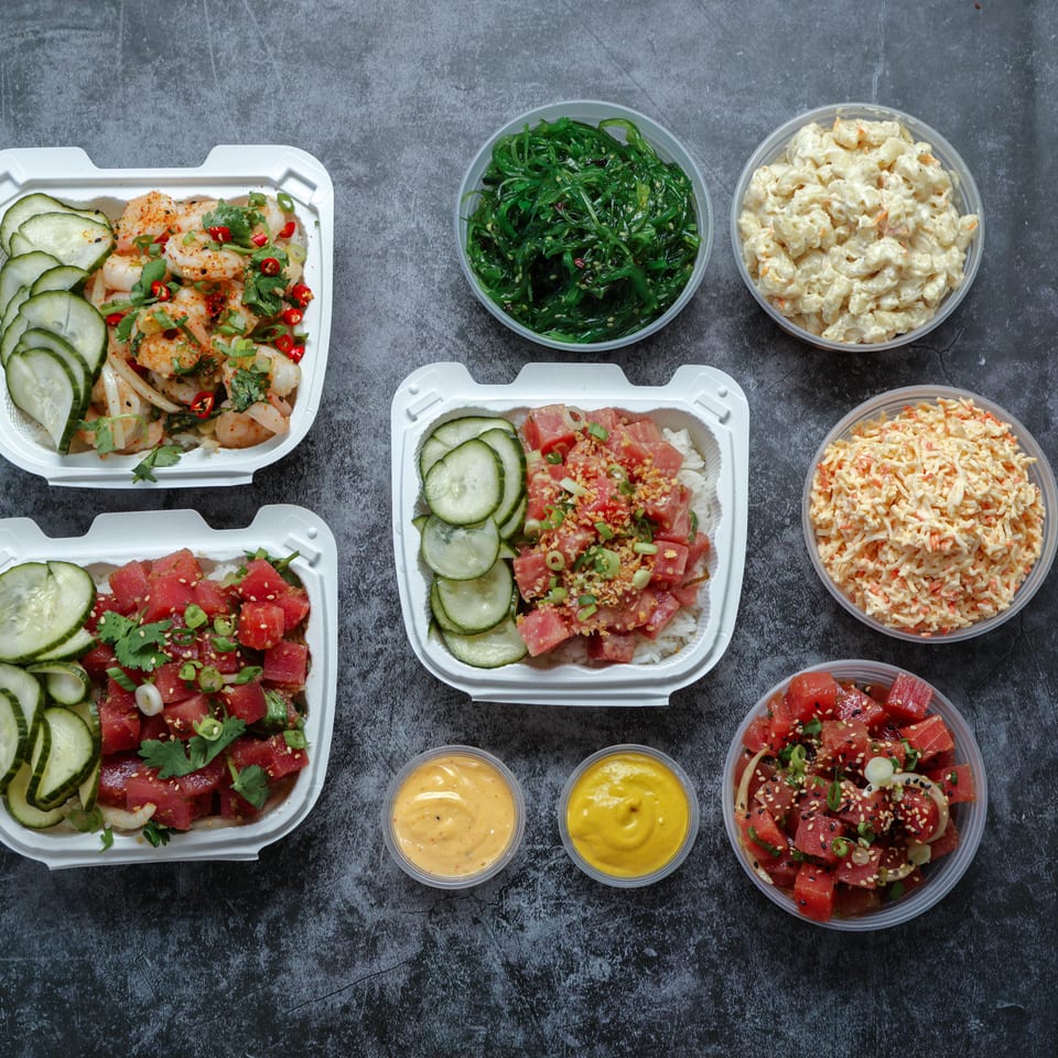 Assorted seafood poke bowls with cucumbers, tuna, shrimp, seaweed salad, creamy sides, sauces, and toppings arranged on a dark tabletop.
