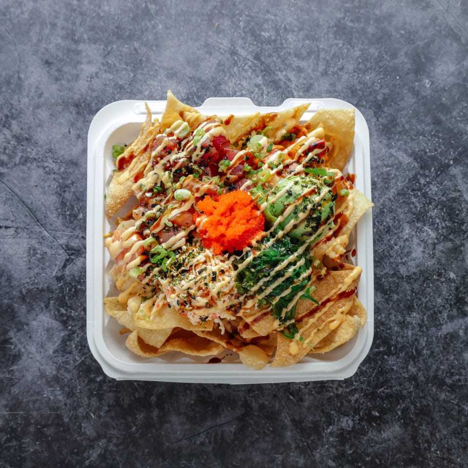 Loaded nachos topped with avocado, shredded vegetables, sauces, sesame seeds, and bright orange roe in a white tray.