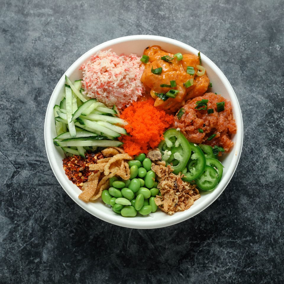 Colorful rice bowl topped with crab, chicken, cucumber, edamame, jalapeños, crispy onions, and chili flakes on a dark stone surface.