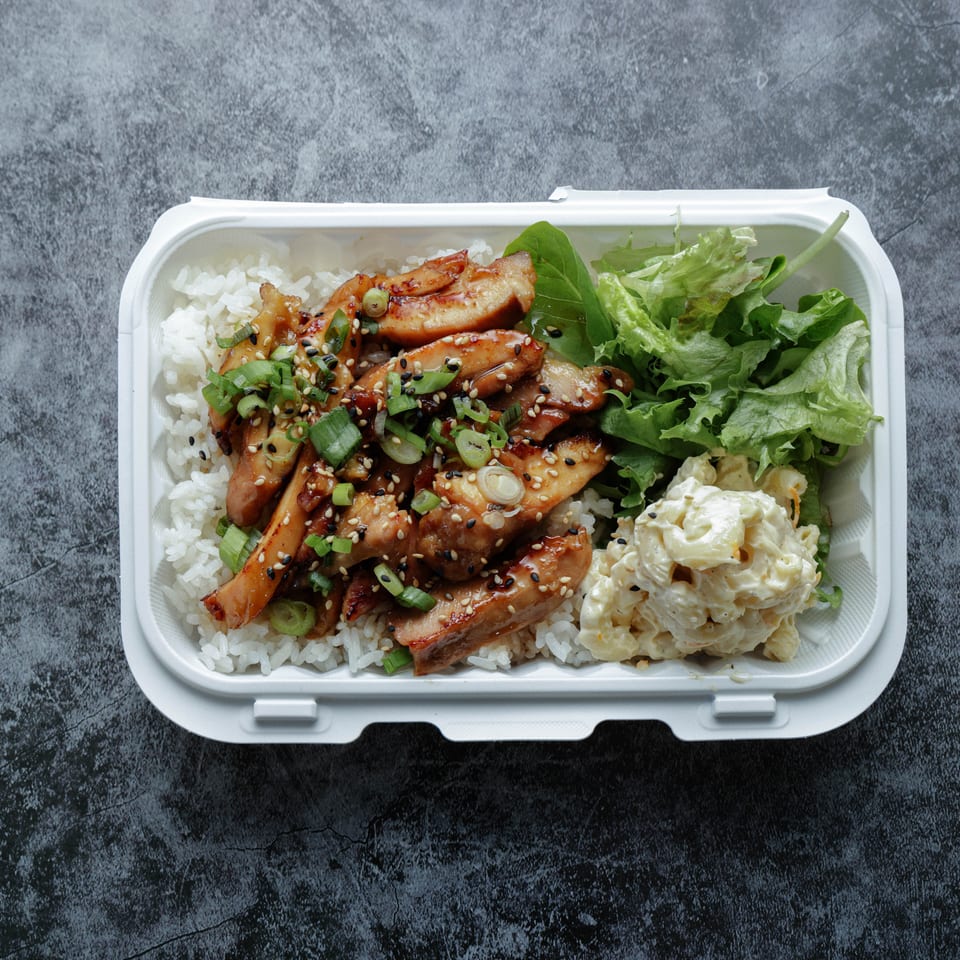 Takeout container filled with white rice, glazed sliced chicken, green onions, leafy salad, and creamy potato salad.