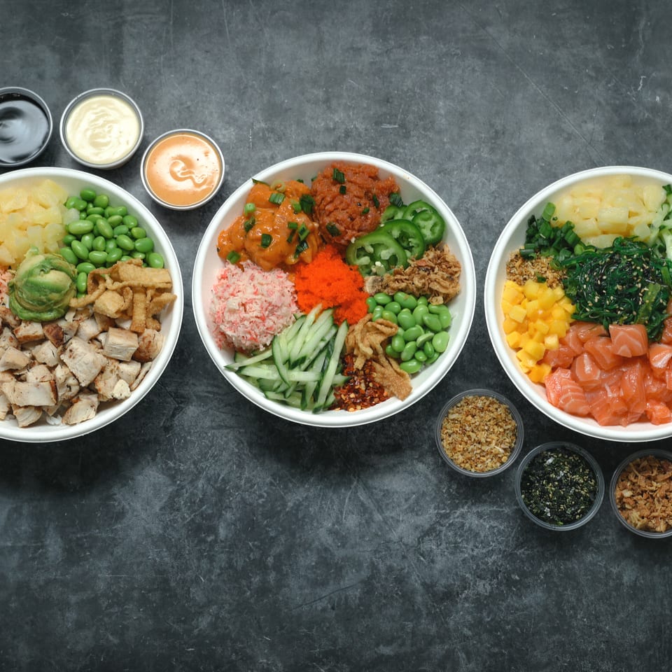 Three colorful poke bowls with salmon, chicken, vegetables, rice, sauces, and toppings arranged on a dark tabletop.
