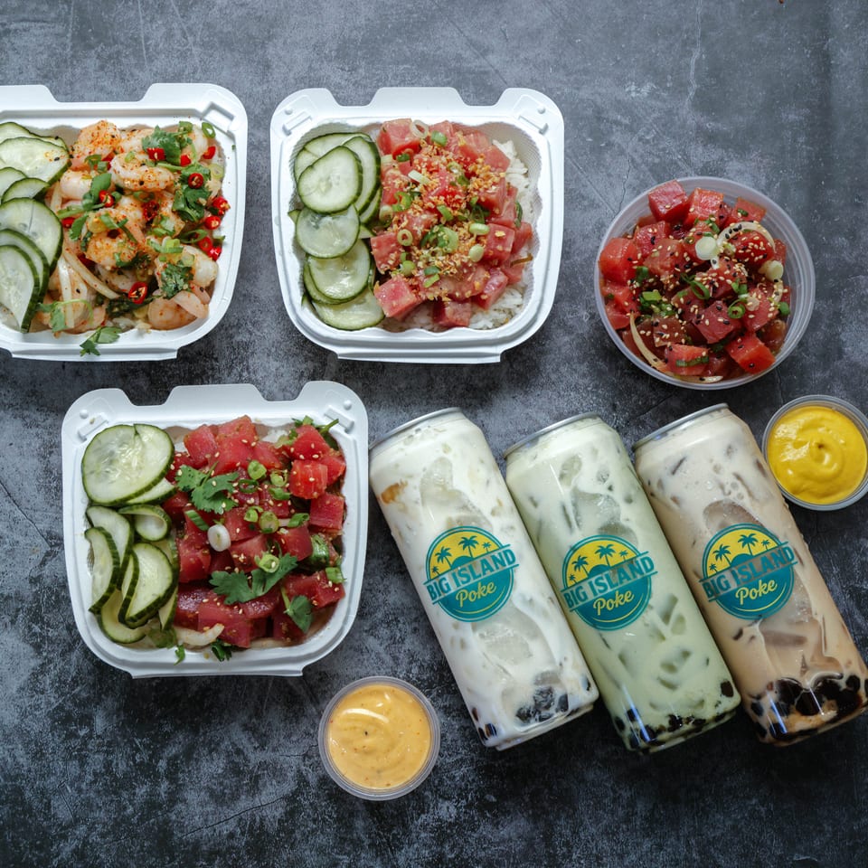 Big Island Poke bowls with shrimp, tuna, and salmon, cucumber garnishes, sauces, and three bottled drinks arranged on a dark tabletop.