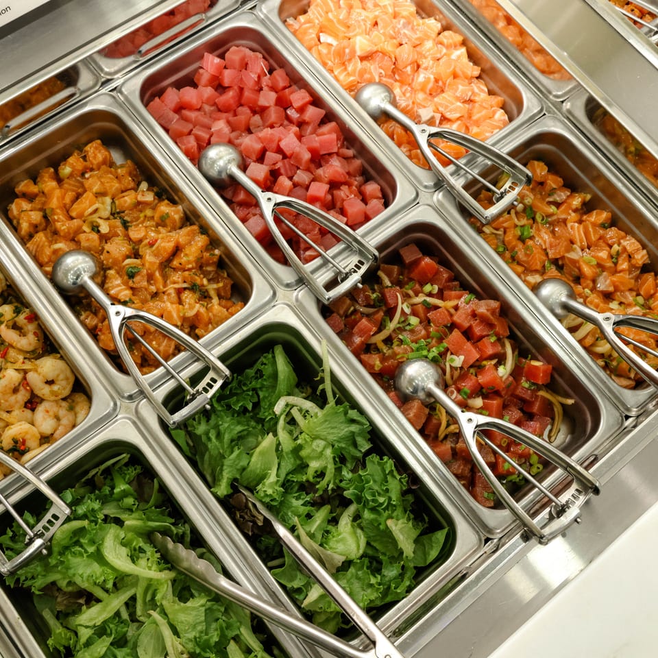 Self-serve buffet displays shrimp, salmon, tuna, mixed seafood, rice, and fresh lettuce in stainless-steel trays.