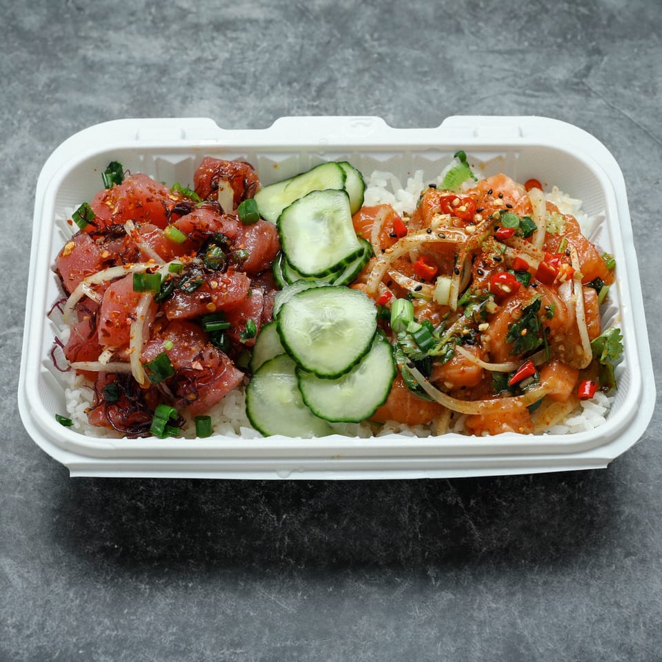 Takeout container of white rice topped with spicy tuna, salmon, cucumber slices, onions, herbs, and red chilies.