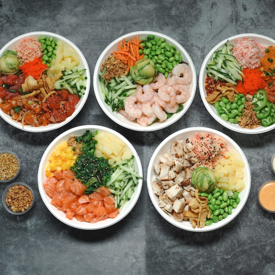 Five colorful poke bowls topped with seafood, chicken, vegetables, edamame, sauces, and garnishes on a dark tabletop.