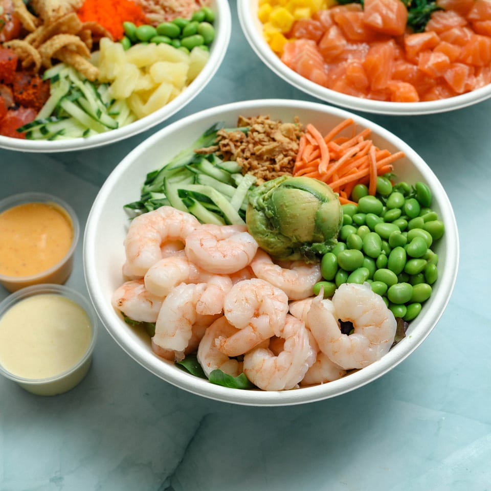 Three poke bowls with shrimp, salmon, vegetables, avocado, edamame, and sauces arranged on a blue marble surface.