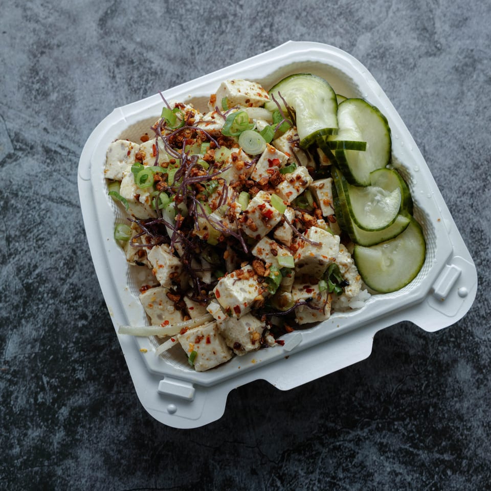 Takeout container filled with seasoned tofu, shredded vegetables, green onions, and sliced cucumbers on a dark stone surface.