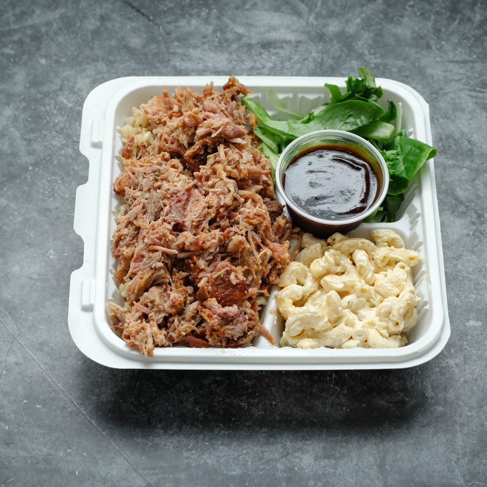 Takeout tray filled with pulled meat, creamy macaroni salad, leafy greens, and a cup of dark barbecue sauce.