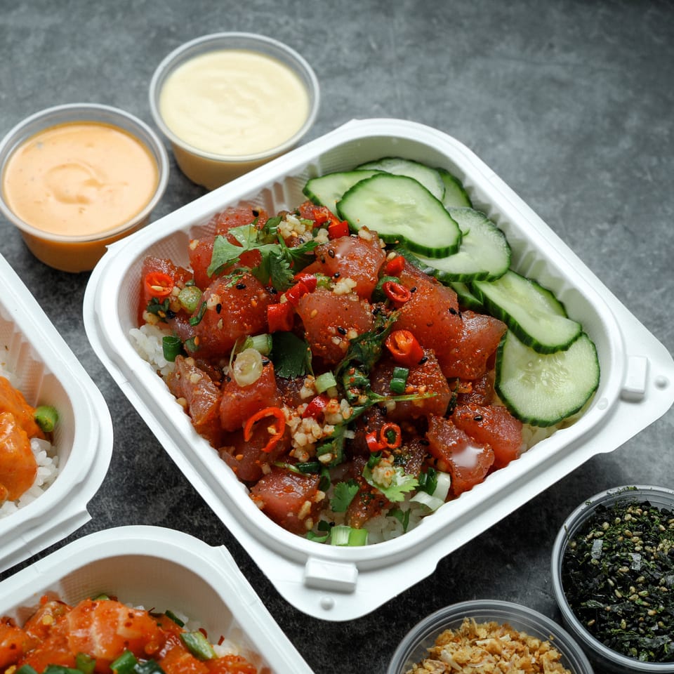 Takeout bowls of spicy tuna poke and creamy orange-topped rice, surrounded by cucumber slices, sauces, and seasoning toppings.