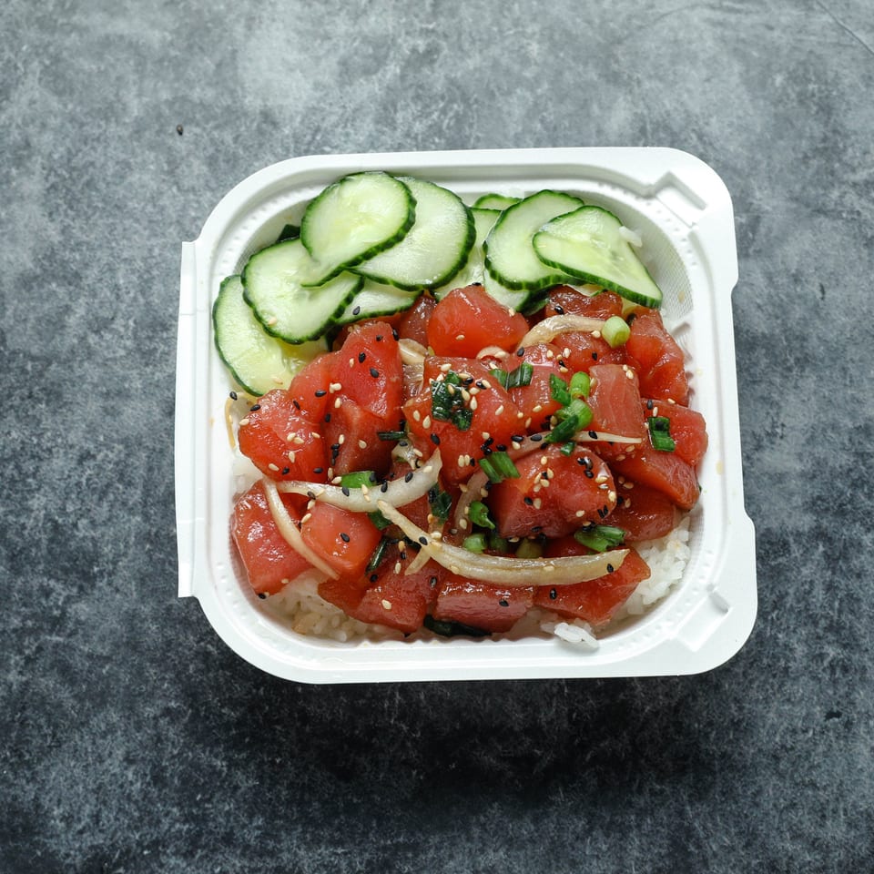 Takeout bowl of diced tuna poke over rice, topped with cucumber, onions, scallions, and sesame seeds.