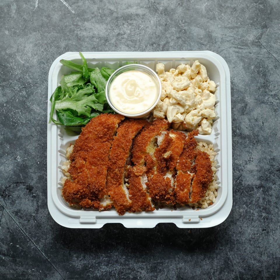 Takeout tray with crispy breaded chicken over rice, macaroni salad, leafy greens, and creamy dipping sauce.